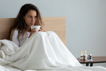 The figure is a photo of a woman appearing ill sitting in bed under a blanket. 