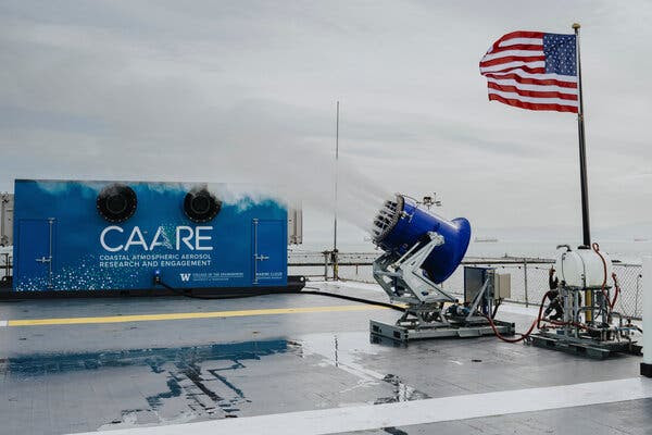 A side view of the spraying machine, looking something like a short cannon, shooting a white mist skyward. The barrel is royal blue. A United States flag waves on a short mast just behind, at the edge of the carrier’s flight deck. 