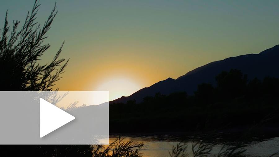 A photograph of a sunset over a mountain and water in the desert with a play button overlay.