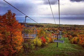 two empty ski lift chairs at the top of a green, grassy slope, the lift cables stretch down through autumn forest with gold, red and orange color
