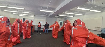 Several people in a classroom with most of them wearing a puffy orange suit