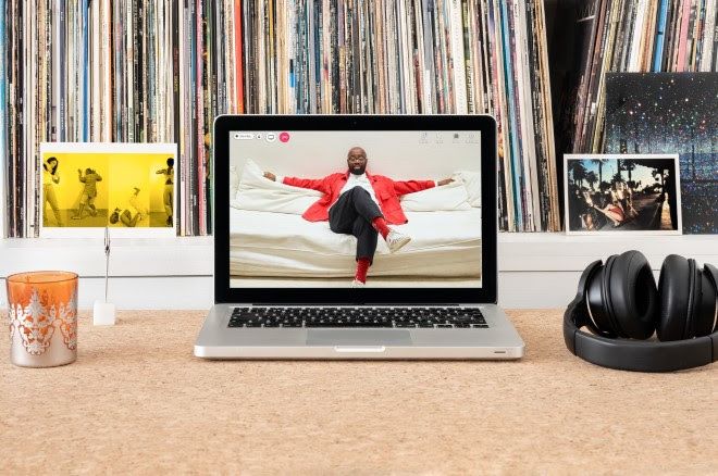Photograph of a laptop on a desk.  On the laptop screen is a portrait of Harold Offeh sitting on a sofa with his arms spread out across the back of the sofa and his legs crossed. Harold is wearing glasses, a red jacket, and a white shirt. Next to the laptop are headphones and a postcard showing a still from Joy Inside Our Tears, four photographs of performers each monotone with a bright yellow tone overlayed.