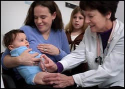The figure is a photograph showing a woman holding a baby, while a clinician administers a vaccine. An older female child is watching.