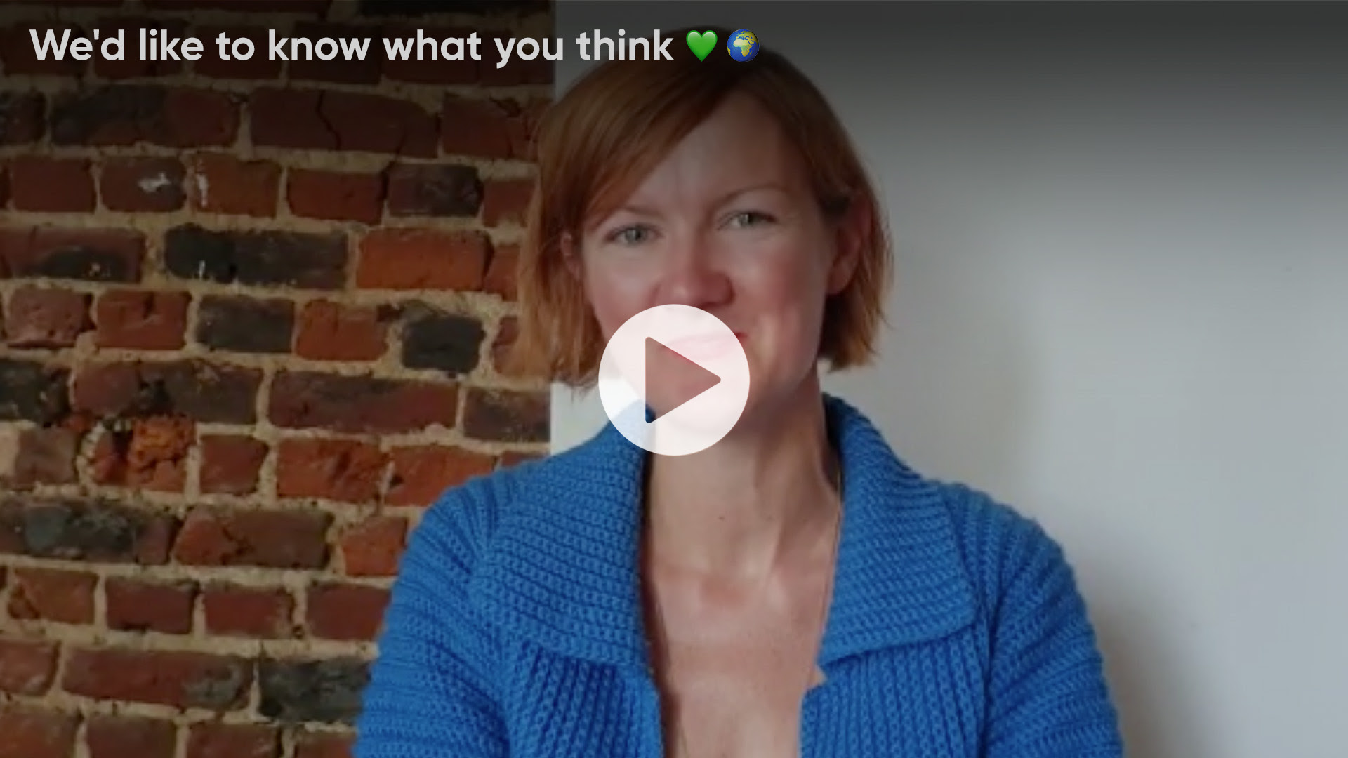 Video titled &quot;We&#39;d like to know what you think&quot;, woman with short red hair and blue top in centre, brick wall behind on left, white on right, play button over top in centre