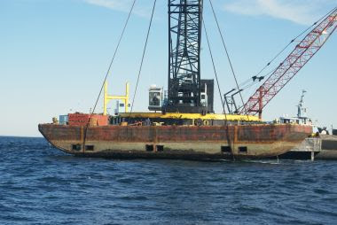 large rusty barge held by crane over ocean surface before being dropped