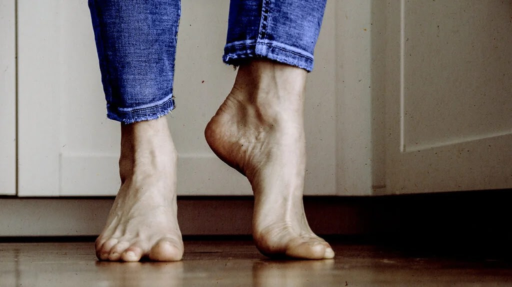 Close-up of someone's bare feet on a wood floor. They are flexing one foot due to the pain of plantar fasciitis.