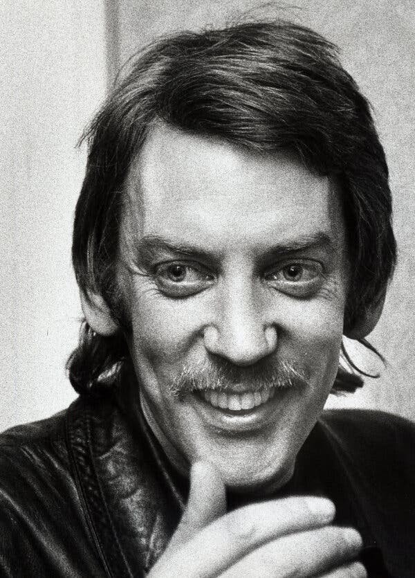 A black and white portrait of a smiling Mr. Sutherland with longish hair and a mustache.