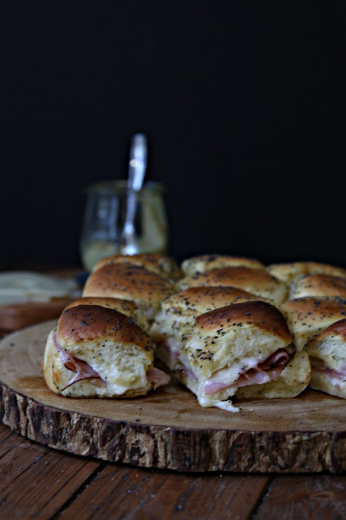 Hot Ham and Cheese Sliders sitting on a decorative wood server. Small glass jar of mustard with spoon in background.