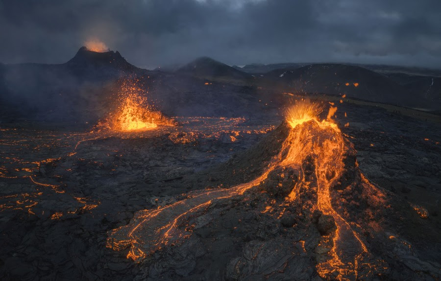 Splashing and flowing lava is seen on a dark lava plain below a volcanic cone, under an overcast sky.