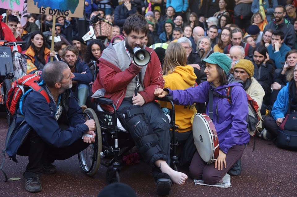 A rebel in a wheelchair, giving a speech with a megaphone. 2 rebels are sat either side of him, with a large group of rebels sat behind them listening to the speech.