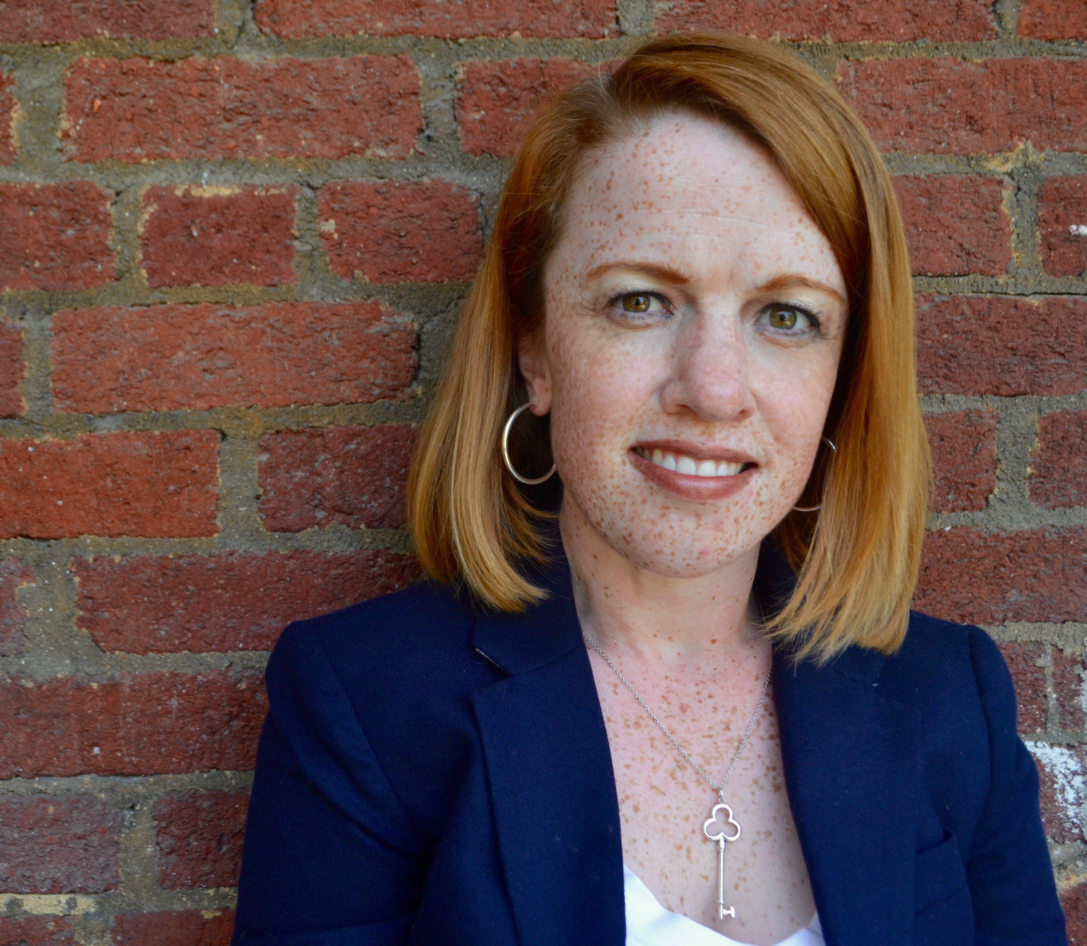 Rebecca Cokley, a red headed woman with a light skin tone smiles at the camera. She is wearing a white blouse, navy blazer, and key necklace. A red brick wall is in the background.