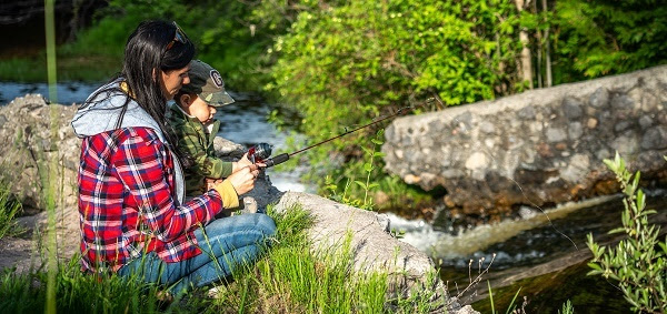 woman helping a young child hold a fishing pole, surrounded by forest greenery and rocks