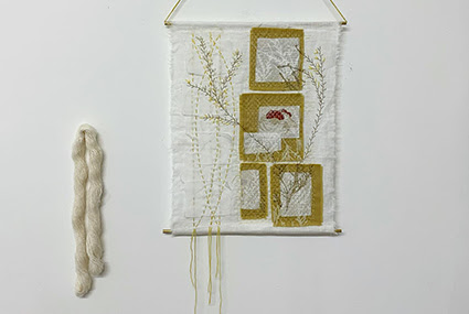 A photograph of a textiles wall hanging by Camban Studio. It is white, with brown and white square details on the right, and floral brown details across. It is displayed on a white wall and next to a strip of white wool.