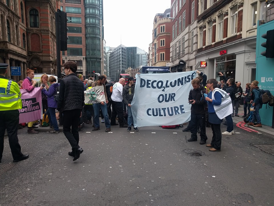 A roadblock on Bishopsgate in London, with around 30 rebels blocking the road. Some activists are holding a banner which reads 'DECO2LONISE OUR CULTURE'.