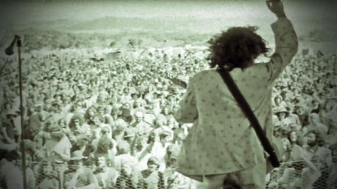 Ross Wilson playing guitar on an outdoor stage with his back to camera, facing a large crowd with his right arm raised, from a screenshot of the film clip for the song Eagle Rock. 
