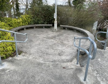 Bottom half of a flagpole with a plaque at its base surrounded by a concrete platform area and steps leading down to this area