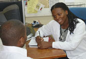 Dr Akwi Asombang in white coat with stethoscope seated at desk takes note on pad while speaking with a seated patient