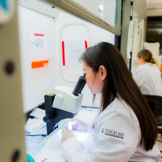 A researcher from the Tisch MS Center of New York peers into a microscope while wearing a white lab coat with the institution's name on the sleeve. Around her, other researchers are working.