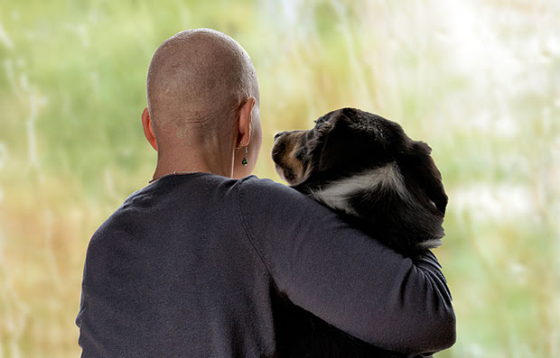 A cancer patient looking out the window with her arm around a dog.