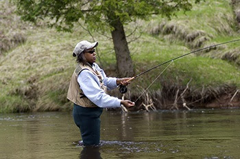 profile view of a woman in dark waders, tan vest and cap, and sunglasses holds a fly-fishing rod, standing thigh-deep in dark green water