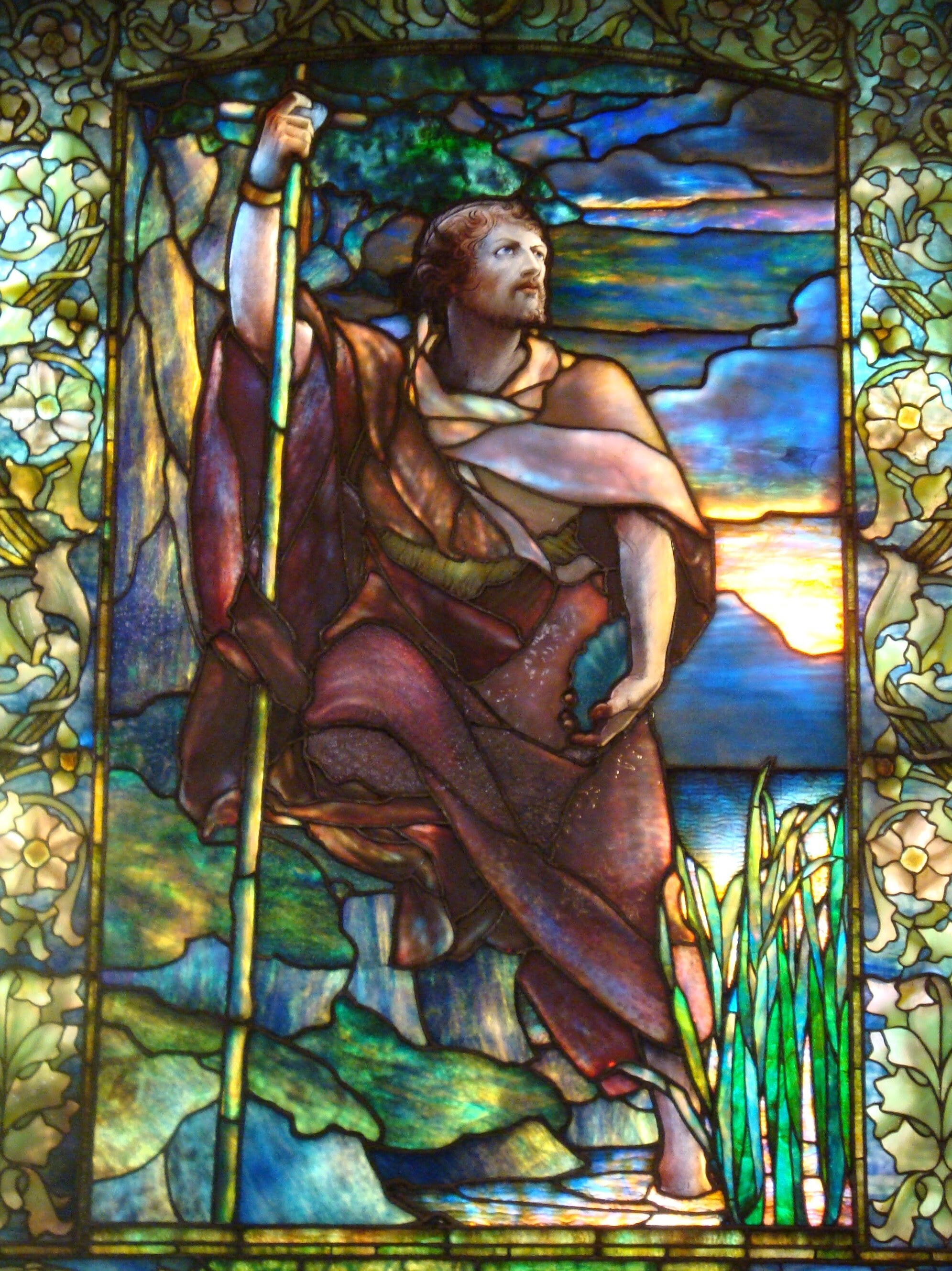 John the Baptist, stands with one foot in the water, ready to baptise people in a stained glass picture by Tiffany.