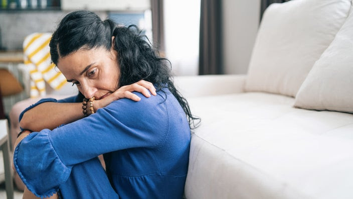 A middle aged woman sitting in front of her couch with her knees pulled into her chest with a sad and dispondent look on her face.