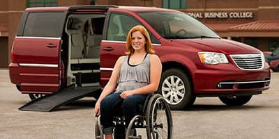A woman in a wheelchair smiles at the camera while sitting in front of an adapted van