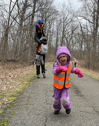 little girl in purple coat and leggings and orange vest smiles while running on paved trail; an adult lifts up a small child in background