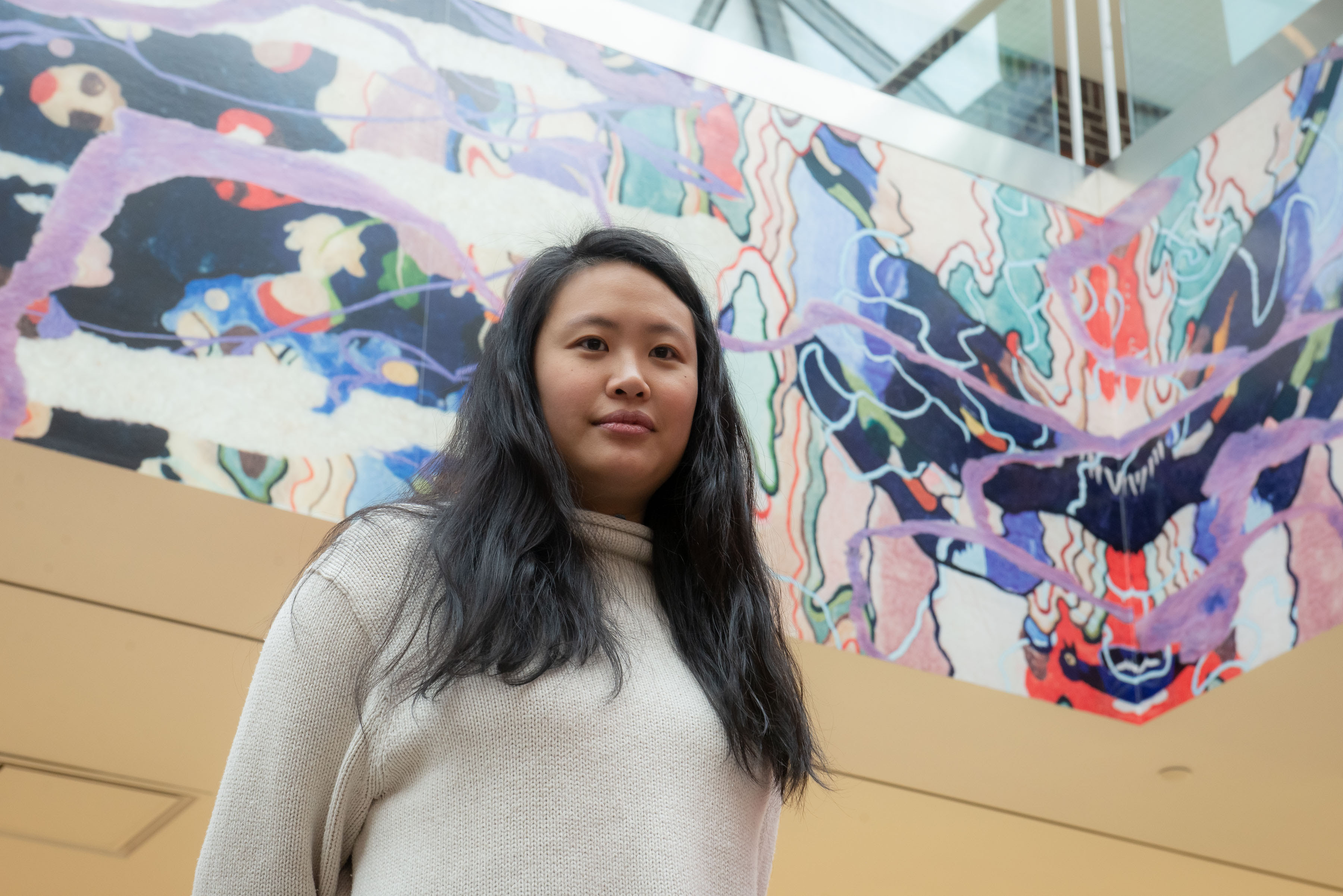 Artist Pauline Shaw is standing in Queens Center Mall with her commissioned mural visible in the background. She has long black hair and is wearing a white turtleneck. Her colorful mural features blue, purple, pink, and pink hues, combining extracted landscapes of birds and flowers with abstracted images of cells and tissues found in the human body.