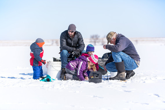 Three young children with their dad and grandpa ice fishing on a frozen lake covered in snow.