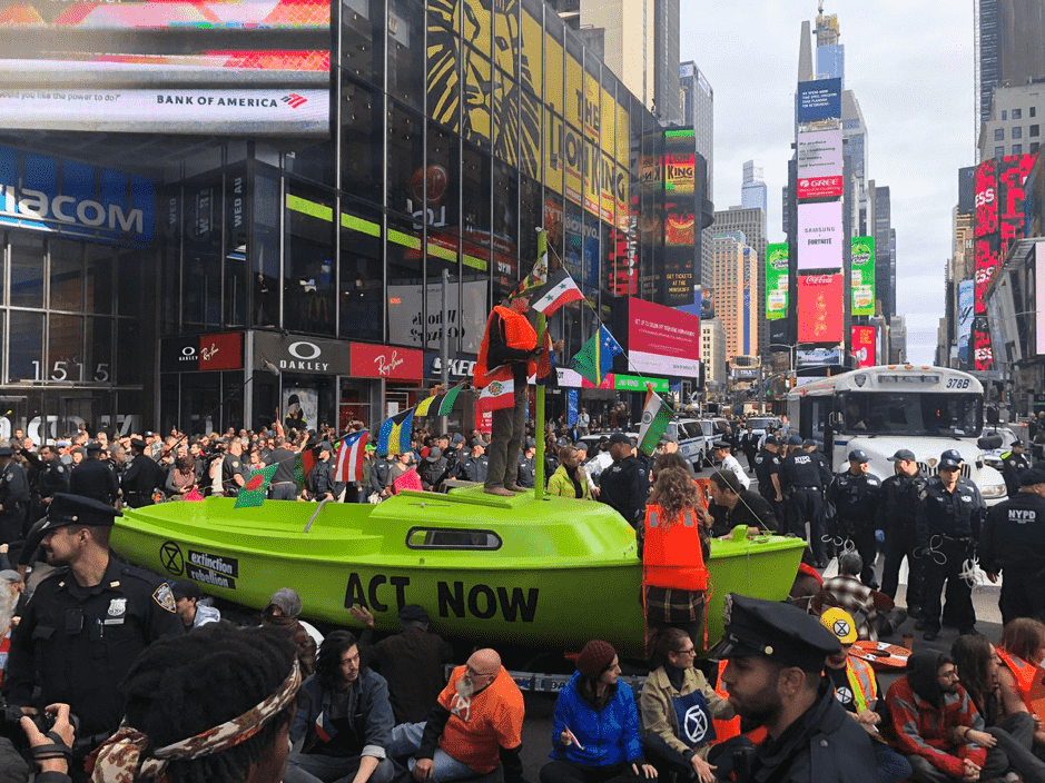 A bright, lime green boat in the middle of Times Square in New York, a very busy intersection of roads. The boat has the words 'ACT NOW' written in black on the side, with lots of police officers surrounding it. A young male activist is stood on top of the boat, which is adorned in the flags of nations which are at immediate risk from sea-level rise.