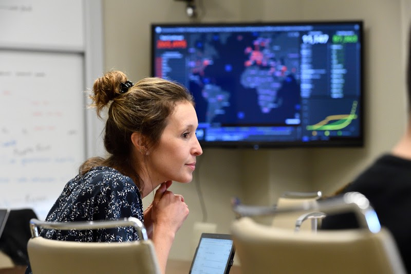 Lauren Gardner sits at a conferece table in front of a monitor showing the Johns Hopkins COVID dashboard