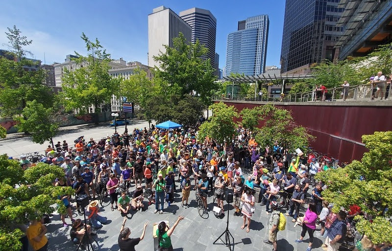 A colorful crowd of people stand in a city plaza surrounded by trees and tall buildings. In the front stands a speaker with a green shirt and long black braid, and an ASL interpreter with a black shirt, both with arms raised to the crowd.