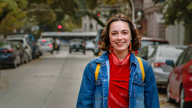A young woman with shoulder-long, curly brown hair stands in the middle of a street. On her right and left are parked cars. She is wearing a red
shirt and a blue jeans jacket and smiles. 