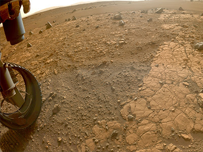 Wide-angle photo from NASA’s Mars Perseverance Rover looks out from underneath the front of the rover at a section of flat, reddish-brown bedrock on a dusty, rocky plain on Mars. Part of the rover’s left front wheel is visible at the lower left.