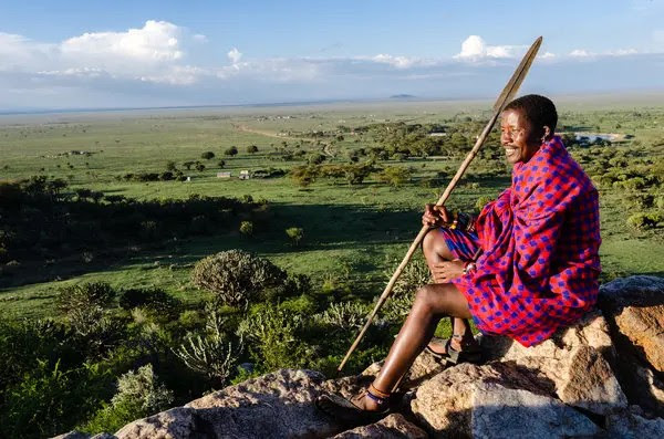 Ein Angehöriger der Massai  sitzt auf einem Felsvorsprung mit Blick auf den üppig-grünen Horizont.
