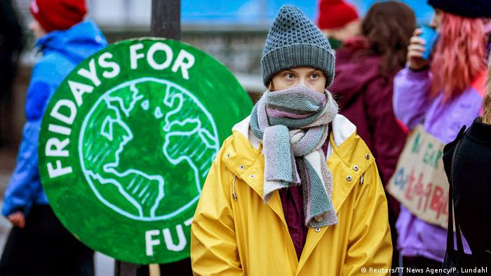 Ativista do clima Greta Thunberg de gorro, jaqueta e rosto meio coberto por cachecol, com cartaz Fridays for Future ao fundo