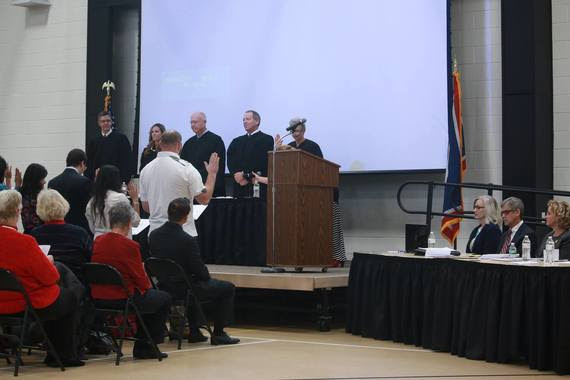 The front row of people in an auditorium stand with their right hand raised to take the Oath of Citizenship before a federal judge.