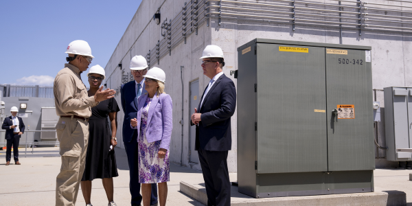 First Lady Jill Biden and Secretary Walsh, wearing hard hats, chat with workers at an electrical substation.