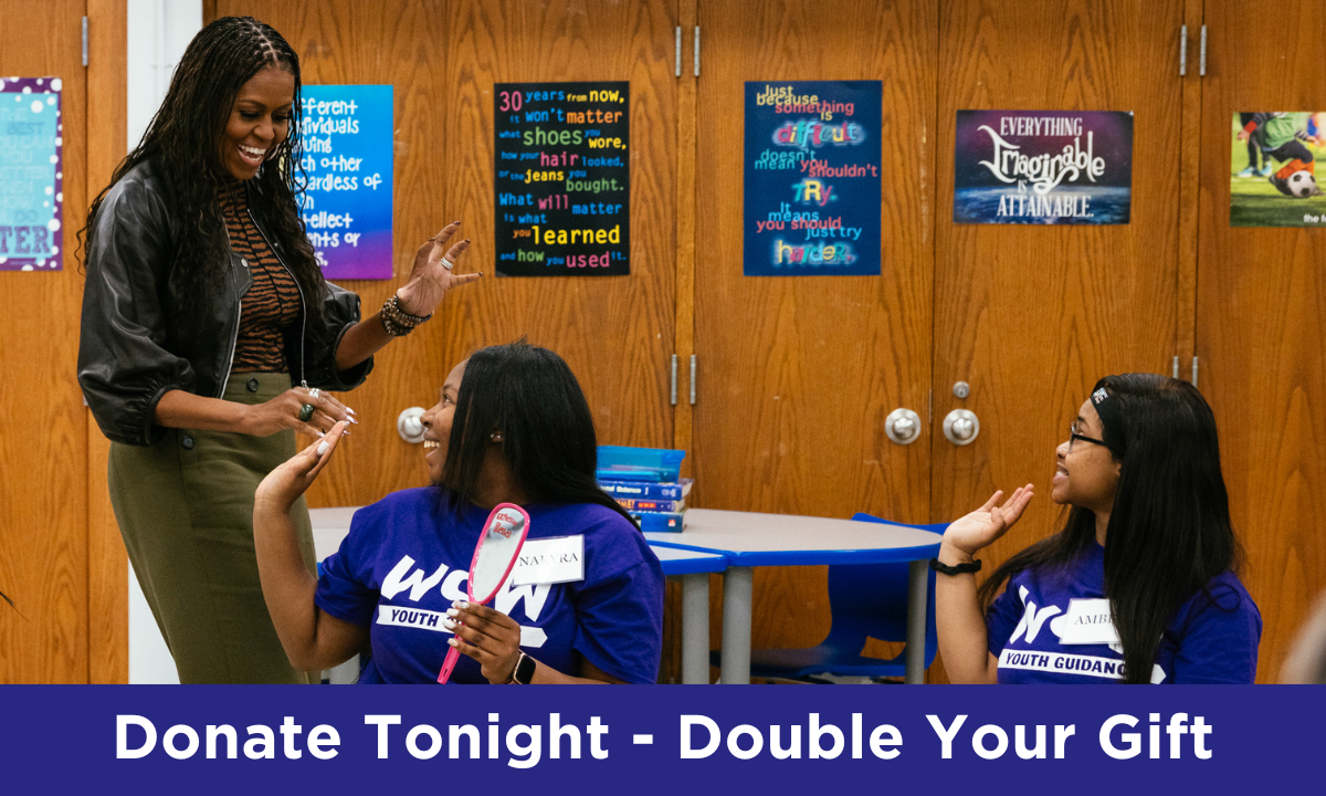 In a classroom setting, Mrs. Obama stands and smiles at two young girls seated in chairs. Both girls have medium deep skin tones and dark hair. Mrs. Obama has her right hand outstretched to greet the girl closest to her. “Donate Tonight – Double your Gift” is written in white text along a blue background on the bottom quarter of the image.